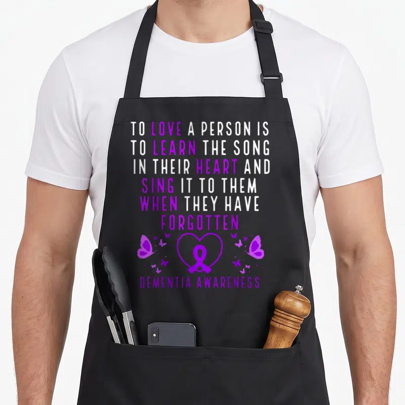 Alzheiemr ribbon apron - Purple and white stacked text with heart ribbon and scattered butterflies on black fabric — shown on a black cotton canvas apron against a white background