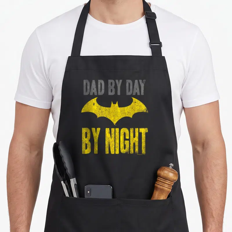 Funny cardinal dad apron - Distressed yellow bat symbol between grey DAD BY DAY and yellow BY NIGHT text — shown on a black cotton canvas apron against a white background