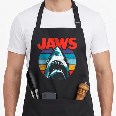 Shark rainbow apron - JAWS text above shark head inside red blue orange white striped circle — shown on a black cotton canvas apron against a white background