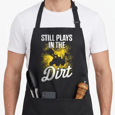 Play heavy canvas aprons - Excavator silhouette with white text Still Plays in the Dirt over yellow splatter — shown on a black cotton canvas apron against a white background