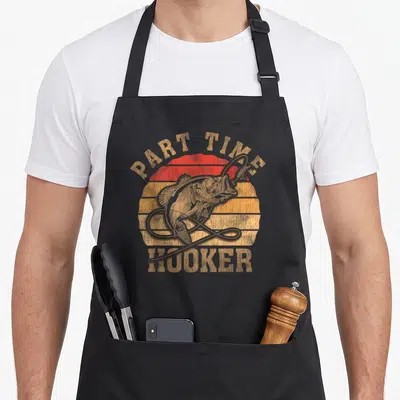 Part dad joke apron - Retro bass fish over striped sunset circle with arched text reading PART TIME HOOKER — shown on a black cotton canvas apron against a white background