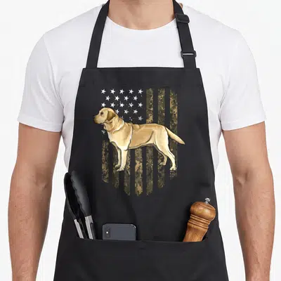 Camo labrador apron - Cartoon yellow lab standing in front of a distressed digital camo flag with stars and stripes — shown on a black cotton canvas apron against a white background