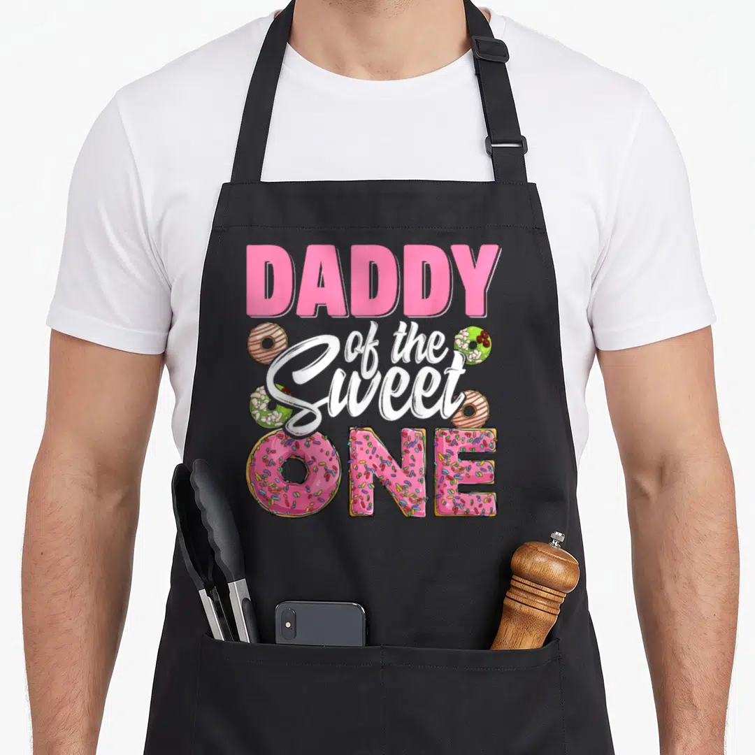 Sweet daddy apron - Pink donut letters spelling ONE surrounded by brown and green frosted donuts with DADDY — shown on a black cotton canvas apron against a white background