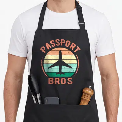 Abroad meme apron - Passport Bros text over striped sun and airplane silhouette in retro colors — shown on a black cotton canvas apron against a white background