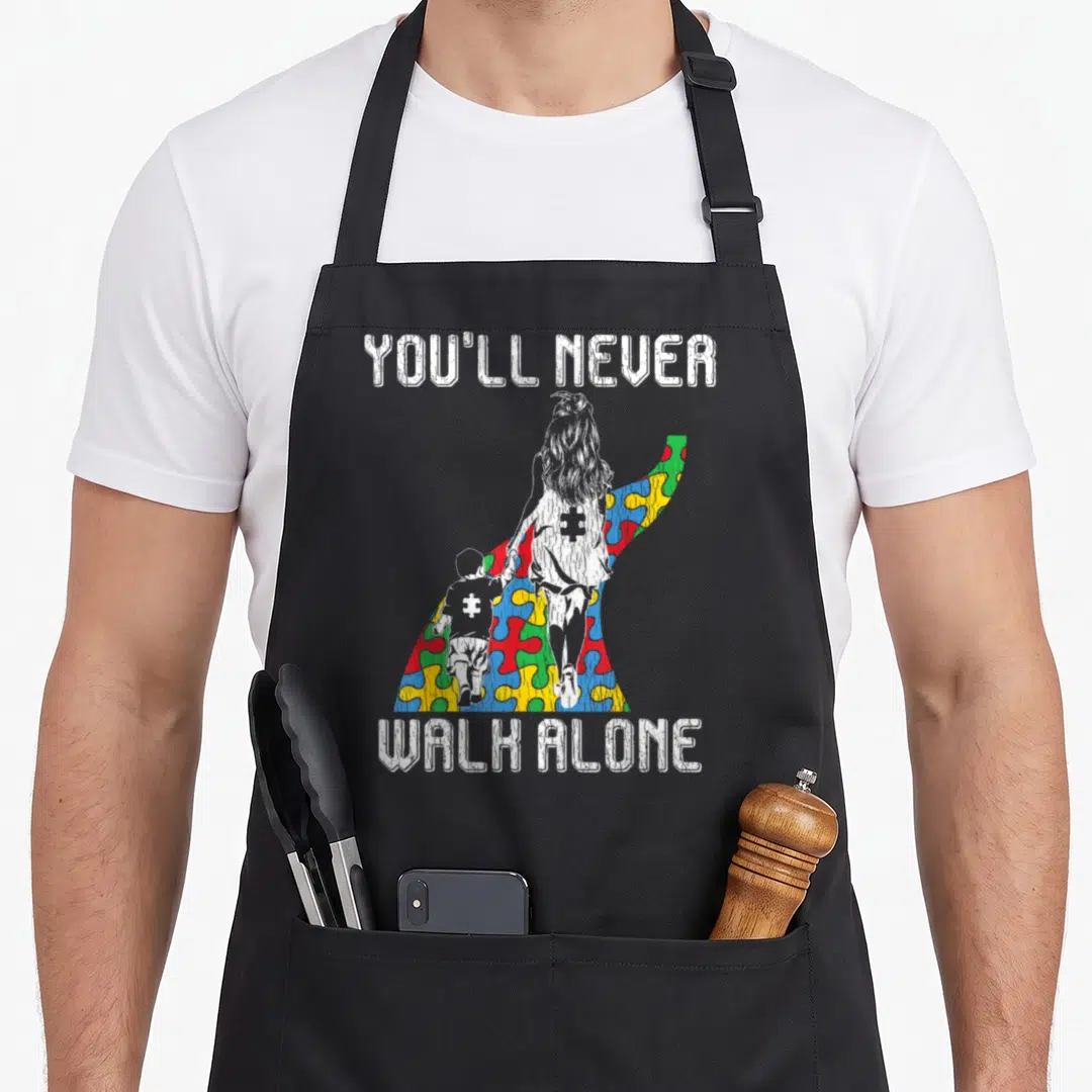 Autism mother son aprons - Distressed text You'll Never Walk Alone above woman and child walking on colorful puzzle — shown on a black cotton canvas apron against a white background