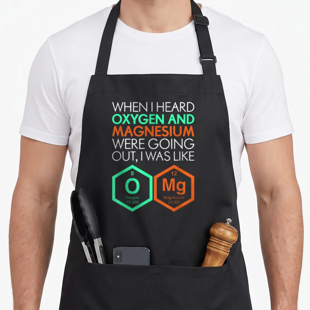 Funny science nerd apron - Stacked text reading When I Heard Oxygen and Magnesium Were Going Out above green and orange — shown on a black cotton canvas apron against a white background
