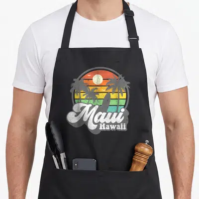 Beach hawaiian apron - Retro sunset circle with palm trees, surfboard, and white Maui Hawaii text — shown on a black cotton canvas apron against a white background