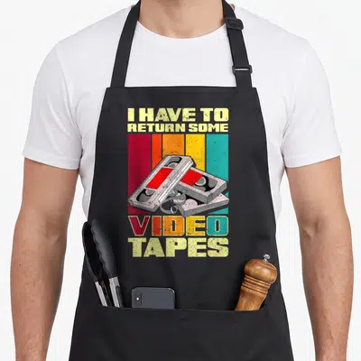 Return 90s apron - I have to return some video tapes text with two stacked VHS cassettes on striped background — shown on a black cotton canvas apron against a white background