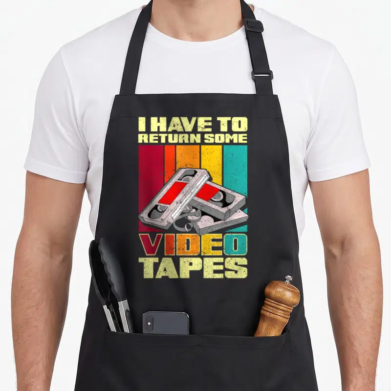 Return 90s apron - I have to return some video tapes text with two stacked VHS cassettes on striped background — shown on a black cotton canvas apron against a white background