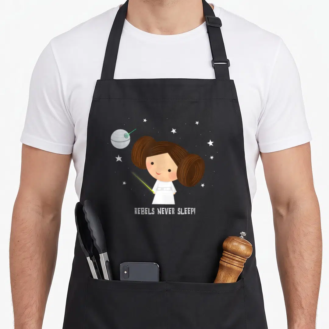 Rebel kawaii apron - Cartoon girl with brown hair buns holding green lightsaber with Death Star and stars above REBELS — shown on a black cotton canvas apron against a white background