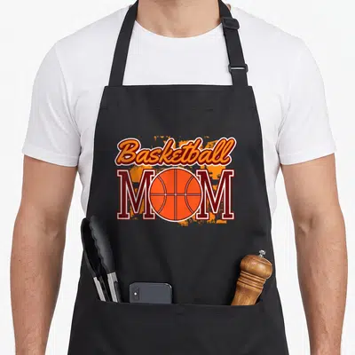 Sport mama apron - Basketball MOM text with orange ball replacing O and splatter background — shown on a black cotton canvas apron against a white background