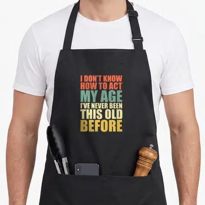 Act year apron - Stacked orange green beige and yellow text reading I don't know how to act my age — shown on a black cotton canvas apron against a white background