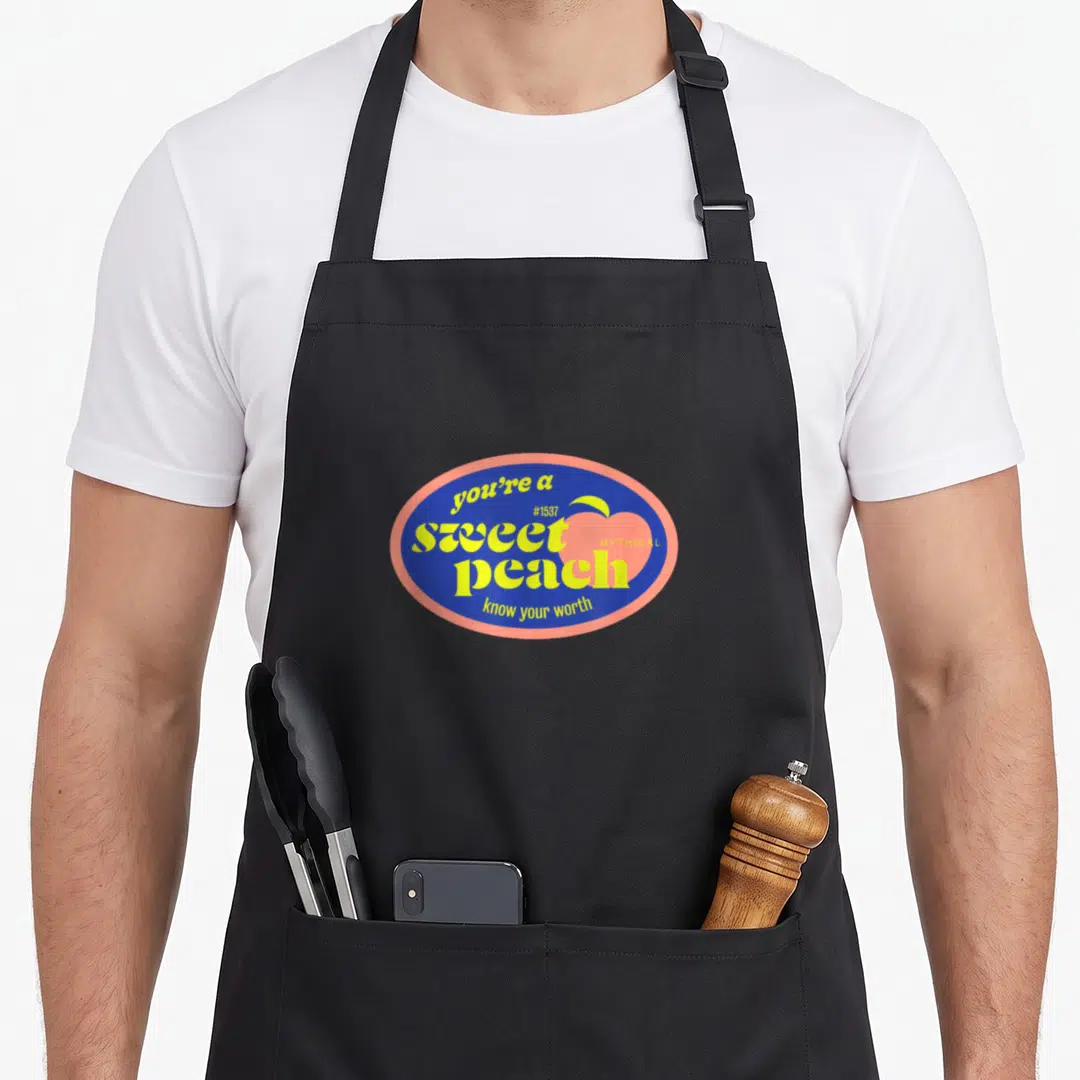 Sweet peach schnapps apron - Blue oval badge with pink peach icon and yellow text reading you're a sweet peach know — shown on a black cotton canvas apron against a white background