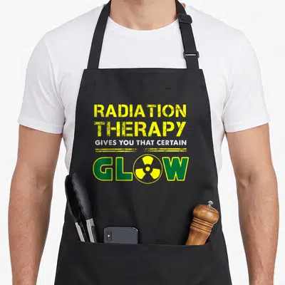 Therapy radiation aprons - Yellow text reading Radiation Therapy Gives You That Certain Glow with a radiation symbol — shown on a black cotton canvas apron against a white background