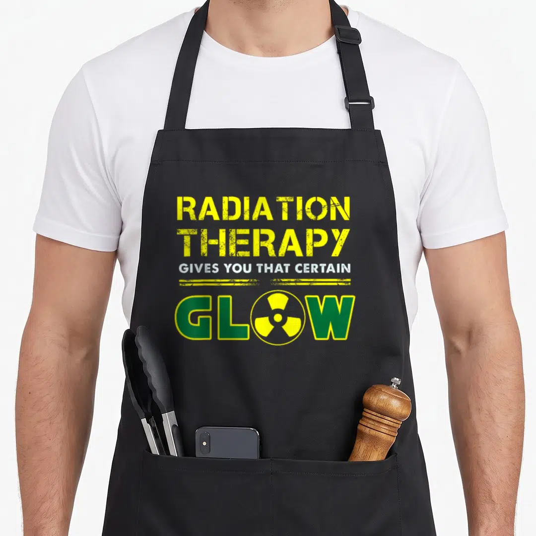 Therapy radiation aprons - Yellow text reading Radiation Therapy Gives You That Certain Glow with a radiation symbol — shown on a black cotton canvas apron against a white background