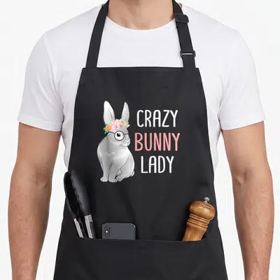 Cute rabbit easter bunny aprons - Grey cartoon bunny with glasses and pink flower crown next to stacked text — shown on a black cotton canvas apron against a white background