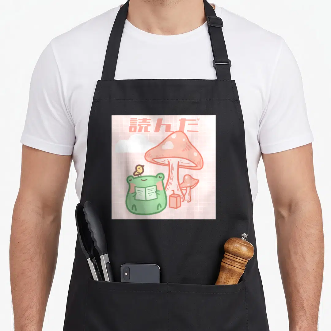 Frog kawaii cottagecore apron - Green frog reading book under pink Japanese text with large mushroom and small bird — shown on a black cotton canvas apron against a white background