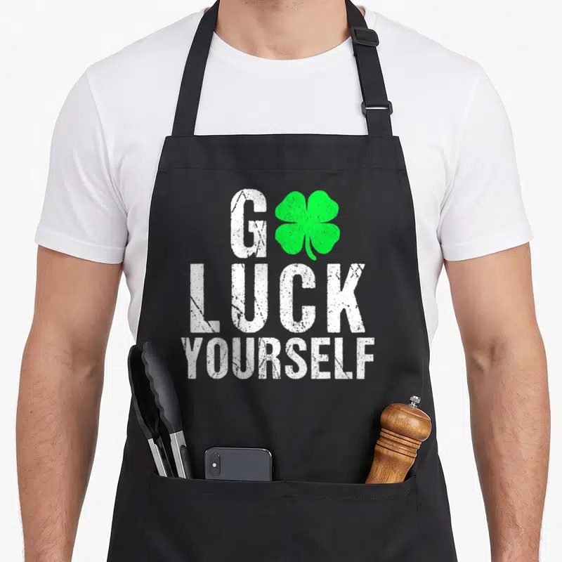 Saint st patricks day apron - Stacked white text Go Luck Yourself with green four-leaf clover replacing O on black — shown on a black cotton canvas apron against a white background