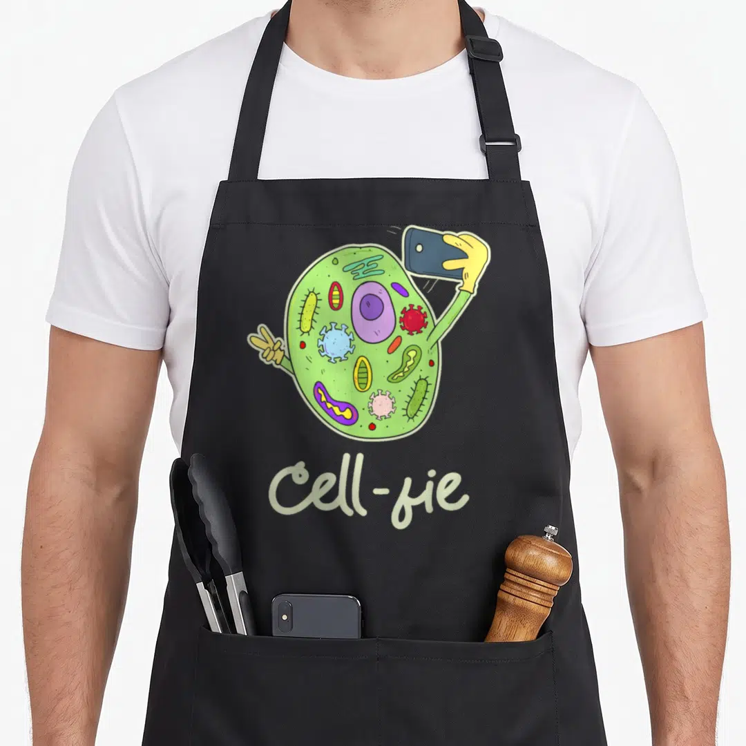 Cell chemistry apron - Green cartoon cell holding smartphone with purple organelles and Cell-fie text — shown on a black cotton canvas apron against a white background
