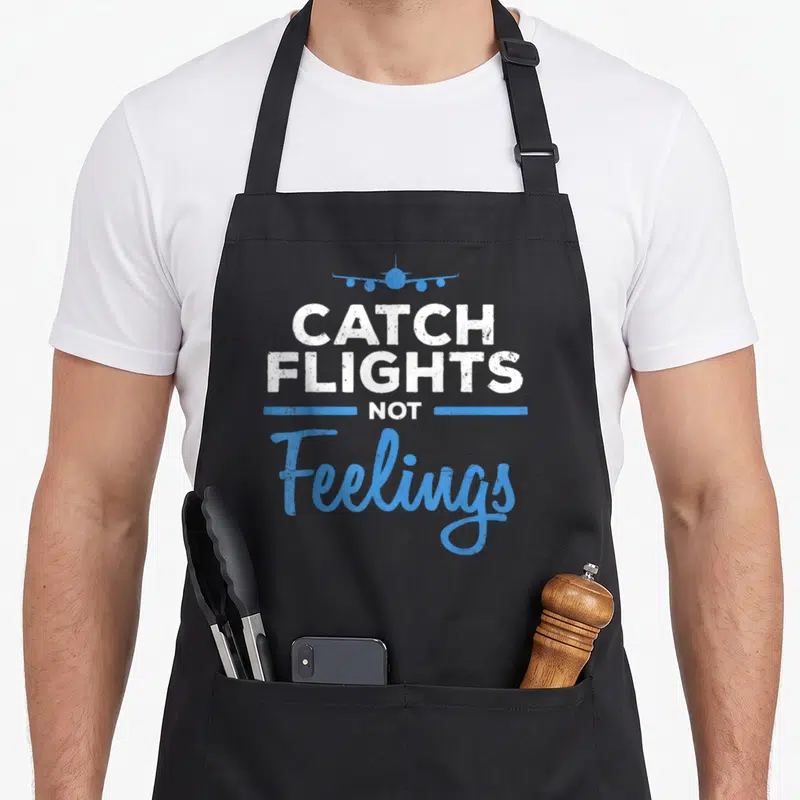Catch flight attendant apron - CATCH FLIGHTS NOT Feelings text with blue airplane icon and horizontal lines — shown on a black cotton canvas apron against a white background