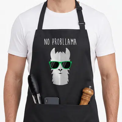 Llama animal apron - NO PROBLLLAMA text above cartoon llama head with green sunglasses — shown on a black cotton canvas apron against a white background
