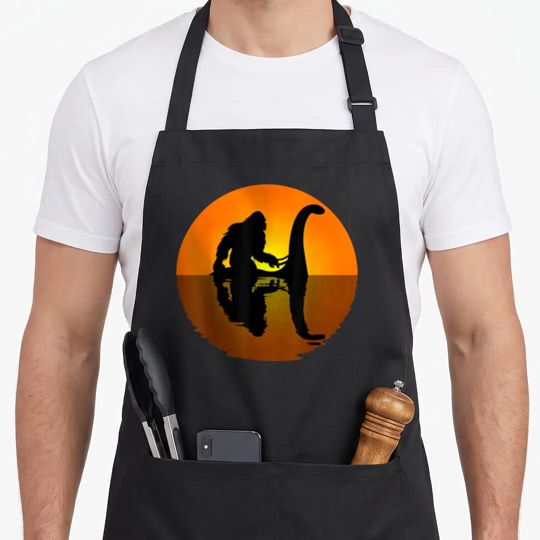 Yeti bigfoot apron - Orange sun silhouette of gorilla riding long-necked aquatic creature with water reflection below — shown on a black cotton canvas apron against a white background