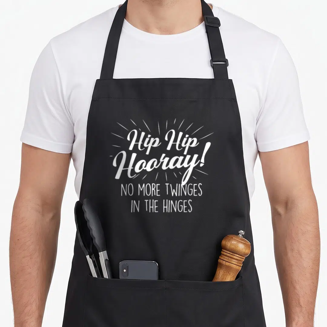 Hooray hip apron - White typography reading Hip Hip Hooray No More Twinges In The Hinges — shown on a black cotton canvas apron against a white background