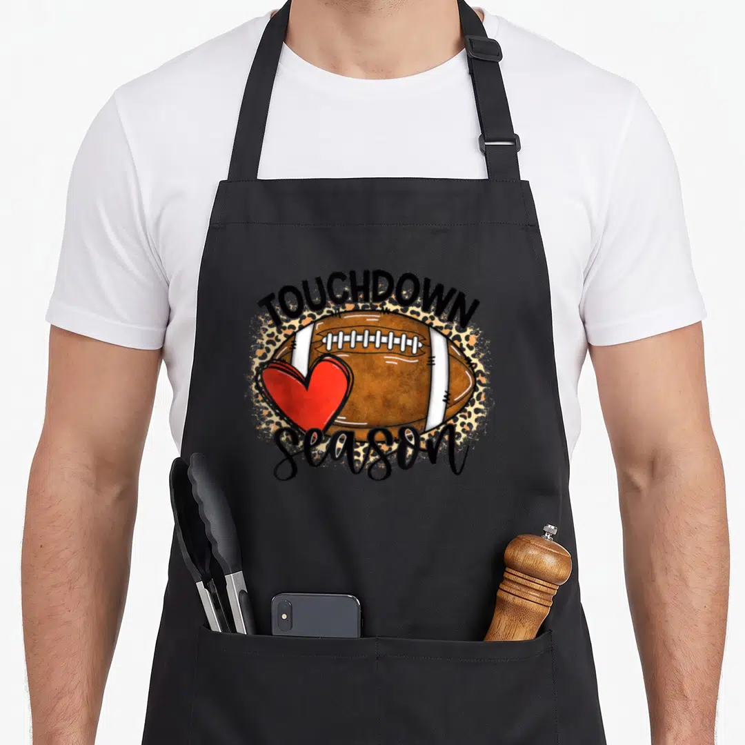 Touchdown leopard apron - Cartoon football with red heart and leopard splatter background reading TOUCHDOWN season — shown on a black cotton canvas apron against a white background