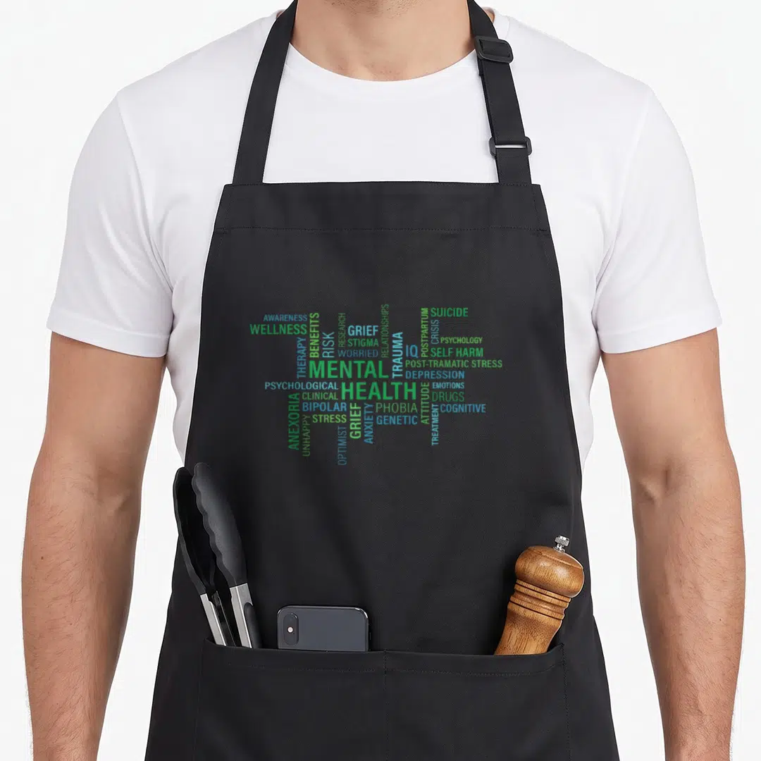 Mental spread apron - Word cloud of mental health terms including depression anxiety therapy and trauma in green blue — shown on a black cotton canvas apron against a white background