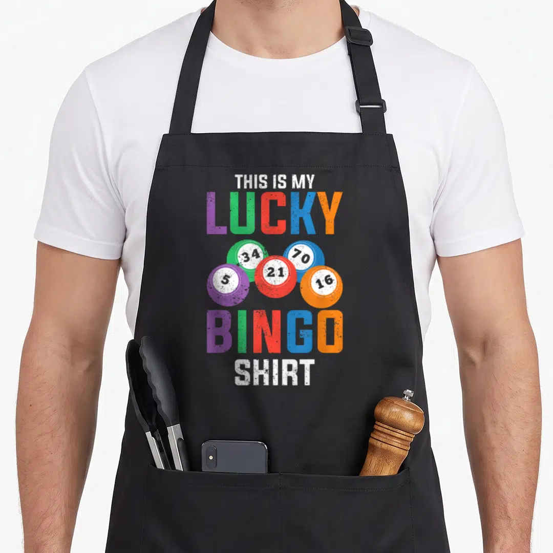 Ist bingo apron - Five numbered bingo balls in white purple green red blue and orange below lucky bingo shirt text — shown on a black cotton canvas apron against a white background
