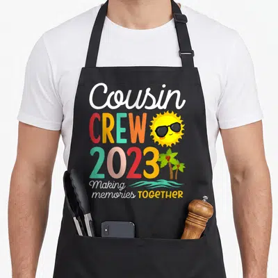 Cousin crew family aprons - Cousin CREW 2023 text with cartoon sun sunglasses palm trees and teal waves — shown on a black cotton canvas apron against a white background