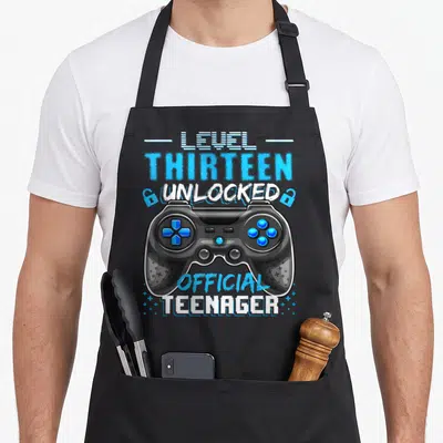 Unlocked 13th video game apron - Blue and white Level Thirteen Unlocked text surrounding a black game controller — shown on a black cotton canvas apron against a white background