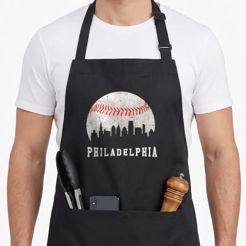 Baseball phillies apron - Distressed white baseball with red stitching and Philadelphia skyline silhouette above text — shown on a black cotton canvas apron against a white background