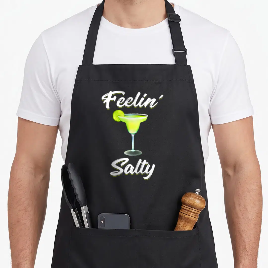 Feelin tommys margarita apron - Cartoon margarita glass with lime slice and white text reading Feelin' Salty on black — shown on a black cotton canvas apron against a white background