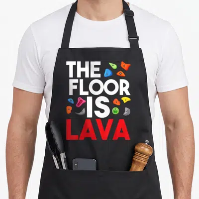Floor bouldering apron - Stacked white and red text THE FLOOR IS LAVA surrounded by blue orange and green climbing holds — shown on a black cotton canvas apron against a white background