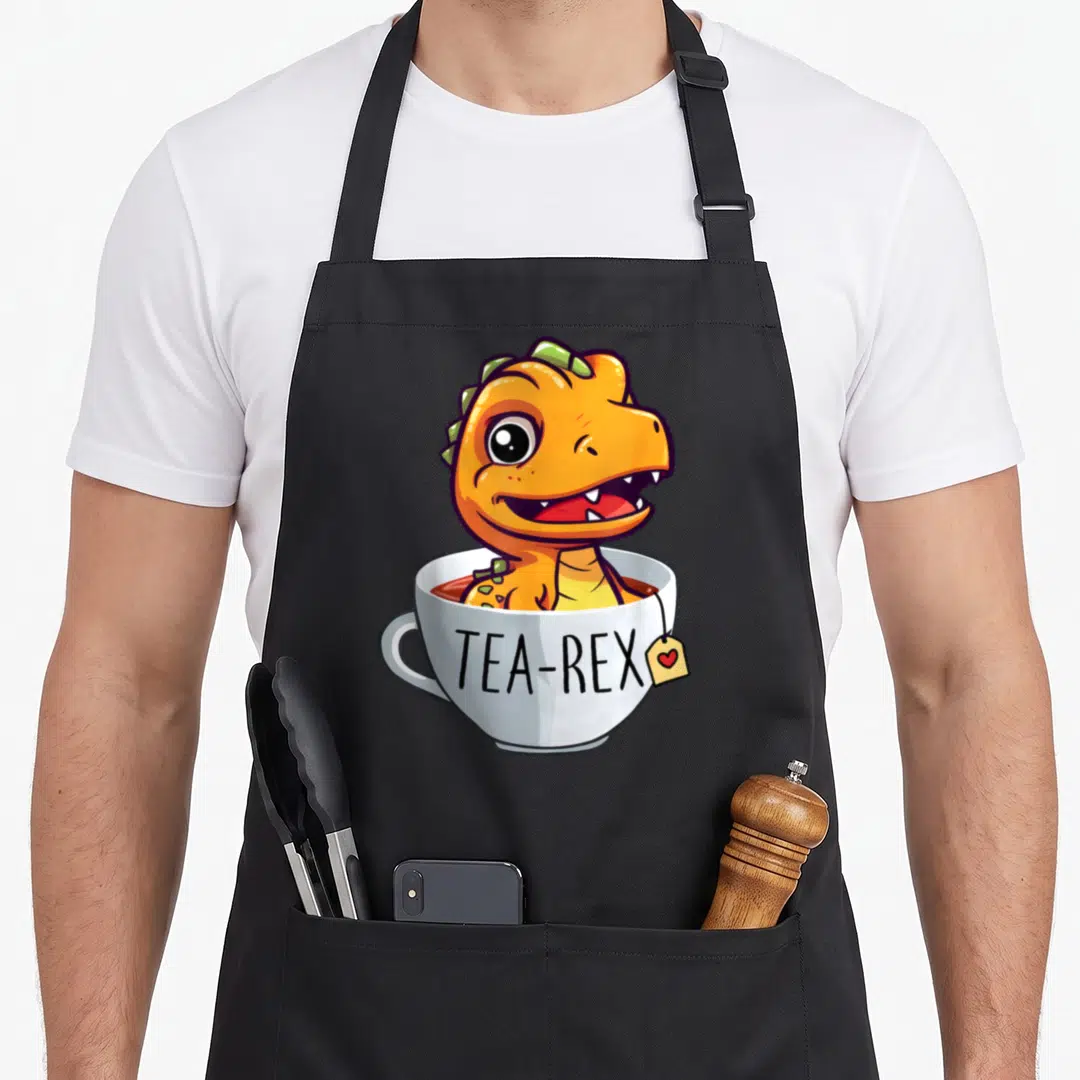 Cup tea party aprons - Orange cartoon dinosaur in white teacup with TEA-REX text and heart tea tag — shown on a black cotton canvas apron against a white background