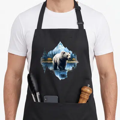 Forest animal bear apron - Realistic bear standing in water with mountain and forest reflection in blue and brown — shown on a black cotton canvas apron against a white background