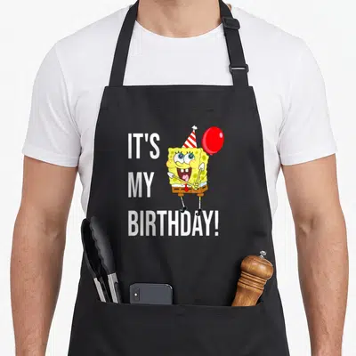 Bday apron - White IT'S MY BIRTHDAY text next to yellow character with red balloon and party hat — shown on a black cotton canvas apron against a white background