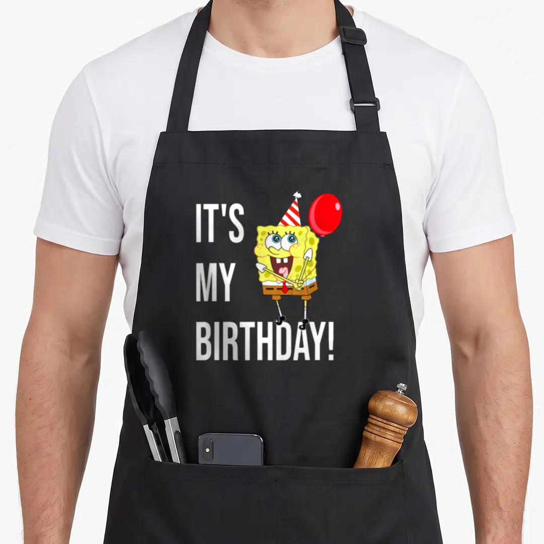 Bday apron - White IT'S MY BIRTHDAY text next to yellow character with red balloon and party hat — shown on a black cotton canvas apron against a white background