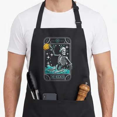 Water skeleton apron - Skeleton in teal water holding orange ball with THE KICKER text inside bordered frame — shown on a black cotton canvas apron against a white background