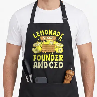 Ceo lemon apron - Cartoon lemonade stand with sliced lemons and arched yellow text reading LEMONADE STAND FOUNDER — shown on a black cotton canvas apron against a white background