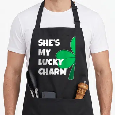 Lucky charm st patrick day aprons - White text She's My Lucky Charm next to a large green shamrock — shown on a black cotton canvas apron against a white background