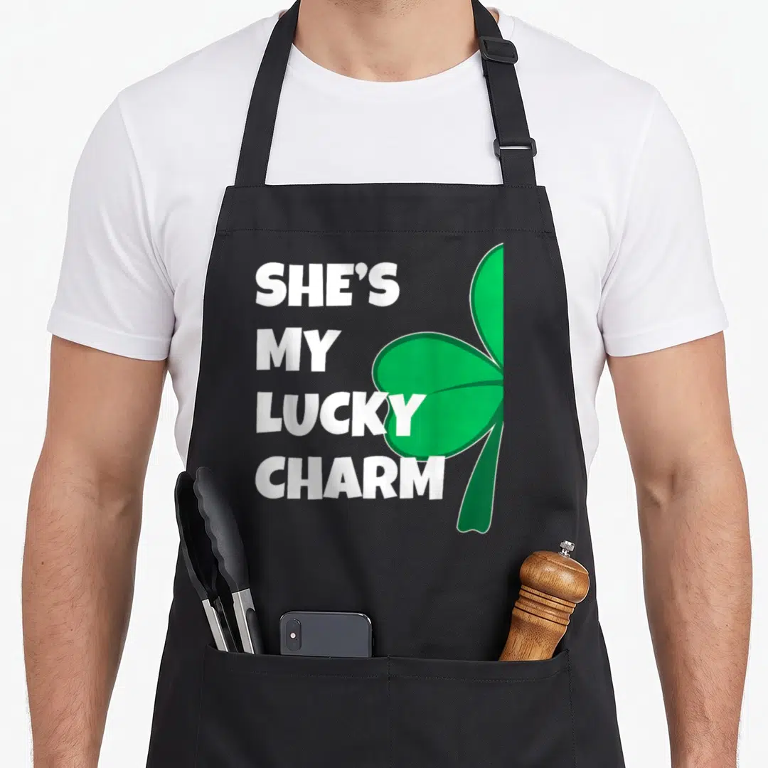 Lucky charm st patrick day aprons - White text She's My Lucky Charm next to a large green shamrock — shown on a black cotton canvas apron against a white background
