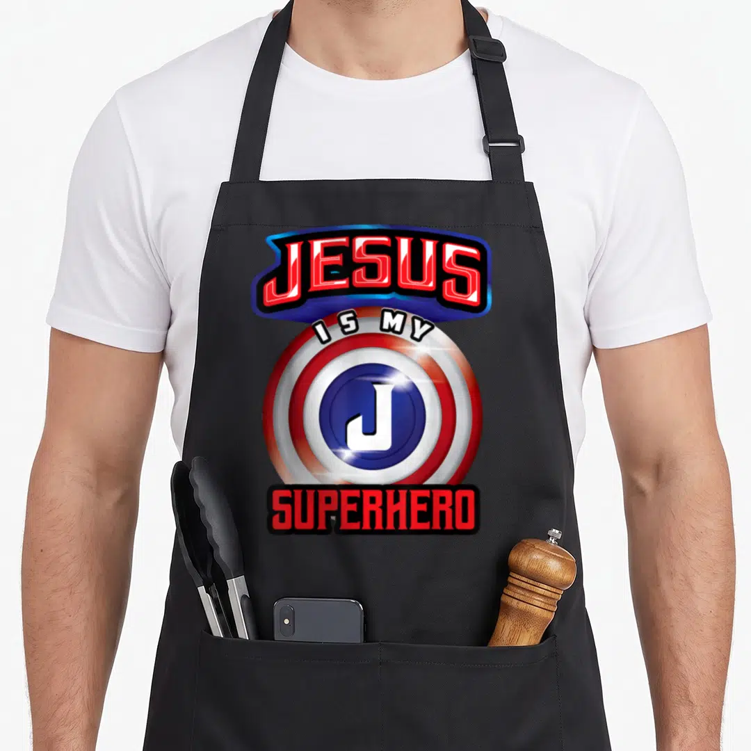 Powerful christian apron - Red white and blue shield with JESUS IS MY SUPERHERO text and white J logo — shown on a black cotton canvas apron against a white background