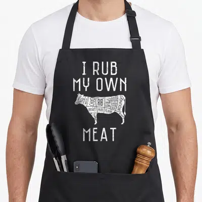 Rub culinary aprons - Distressed white text I Rub My Own Meat above a segmented black cow diagram showing beef cuts — shown on a black cotton canvas apron against a white background