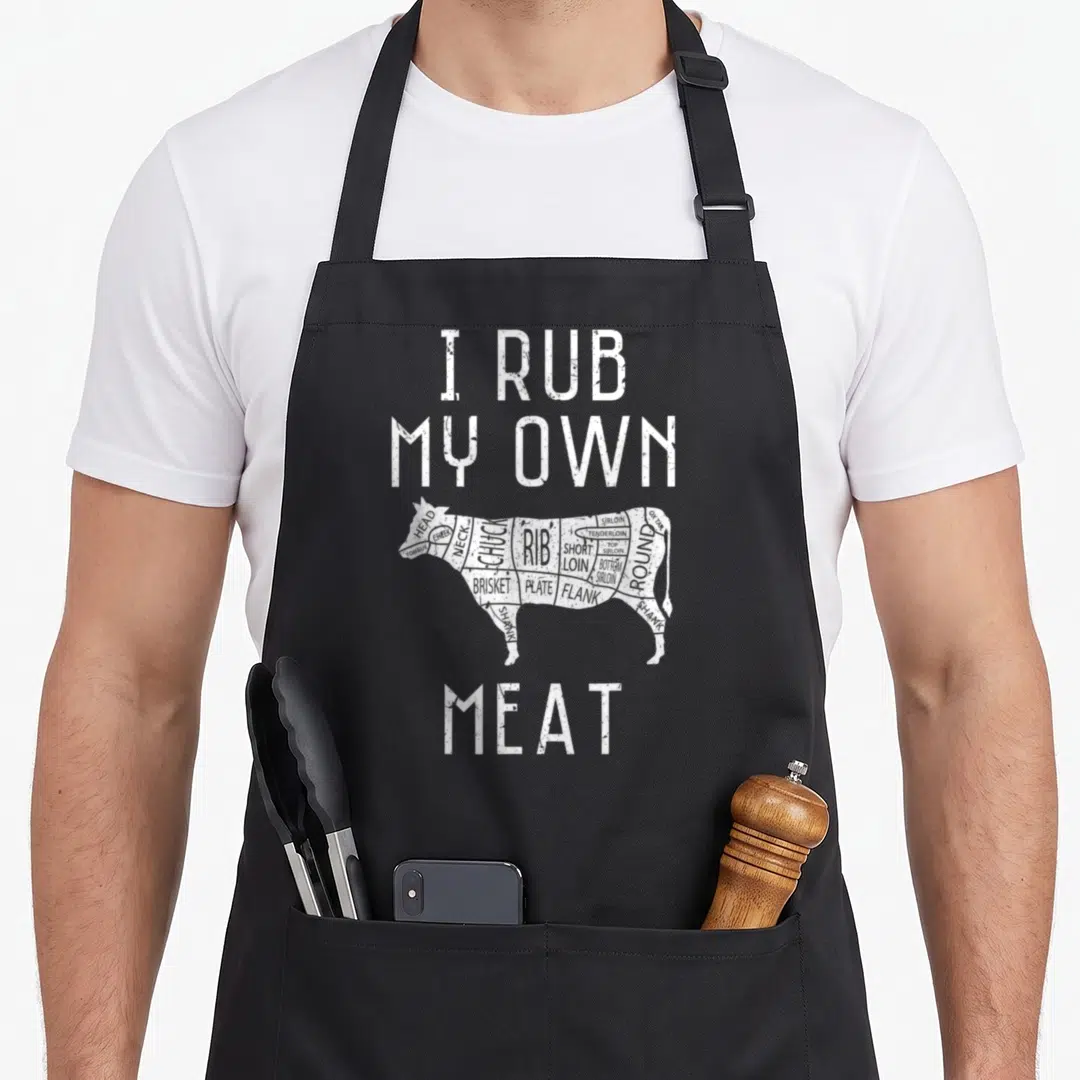 Rub culinary aprons - Distressed white text I Rub My Own Meat above a segmented black cow diagram showing beef cuts — shown on a black cotton canvas apron against a white background