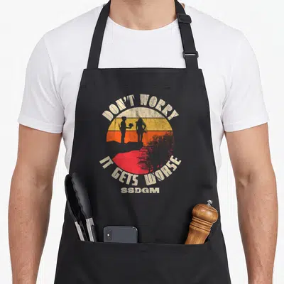 Worry funny key apron - Retro sunset with two silhouettes and text DON'T WORRY IT GETS WORSSDGM in beige orange red — shown on a black cotton canvas apron against a white background