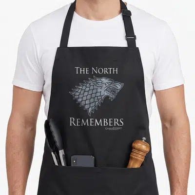 Direwolf north apron - Light grey wolf head line art between THE NORTH and REMEMBERS text — shown on a black cotton canvas apron against a white background