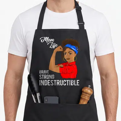 Afro messy aprons - Cartoon woman flexing arm with text Mom Life BRAVE STRONG INDESTRUCTIBLE in red blue white and brown — shown on a black cotton canvas apron against a white background
