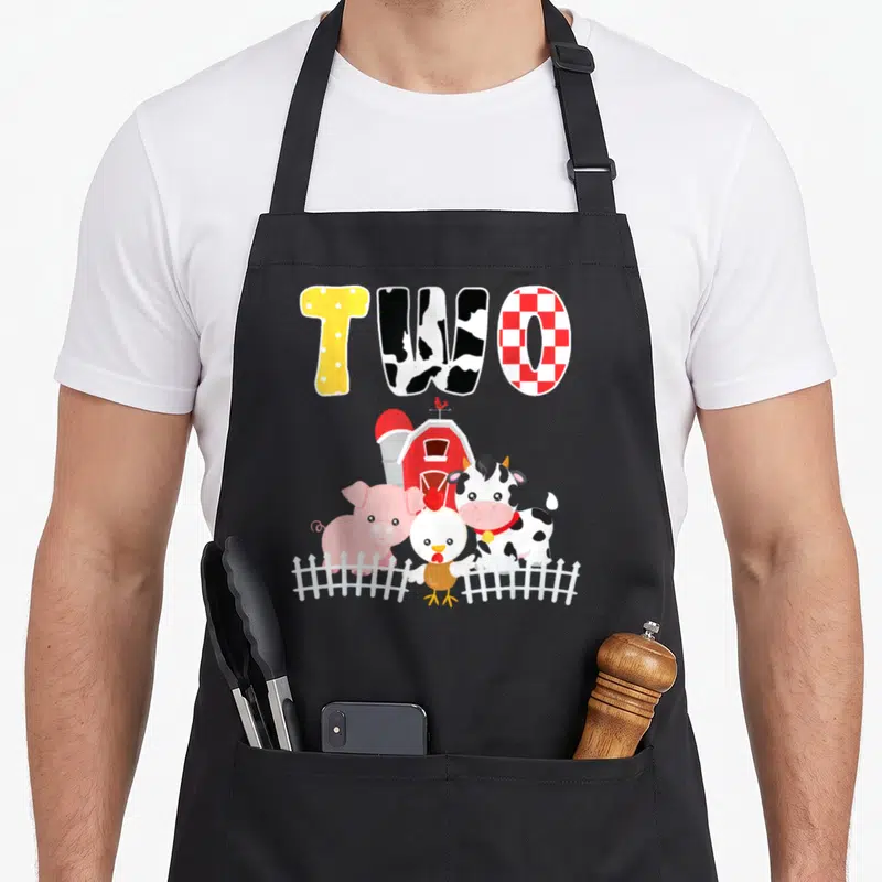 Barnyard animal farm aprons - Cartoon cow pig chicken behind white fence under patterned word TWO — shown on a black cotton canvas apron against a white background